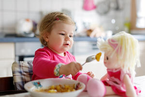 Adorable baby girl eating from fork vegetables and pasta. Little child feeding and playing with toy doll